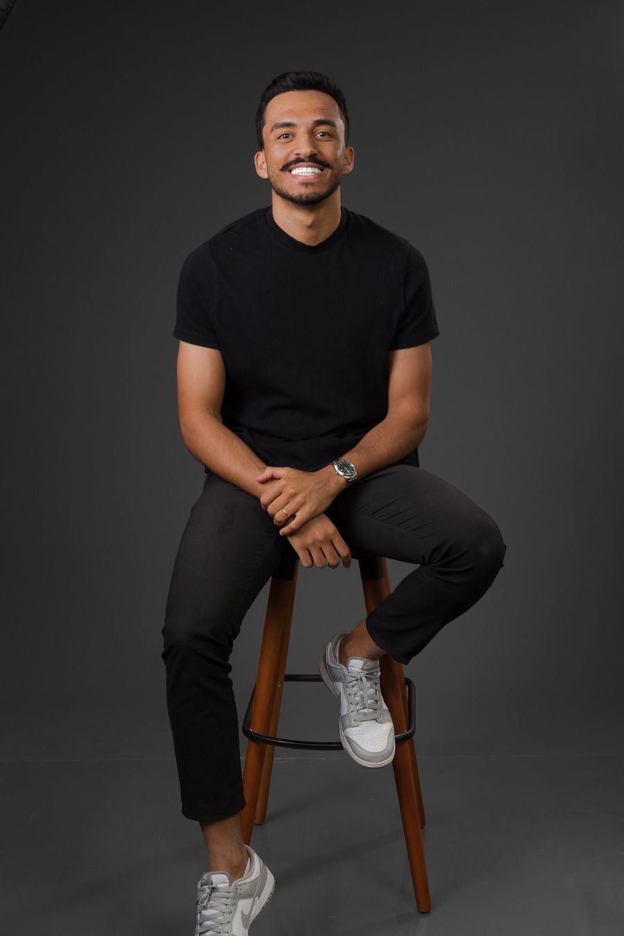 Thiago Nascimento sentado em um banco alto, camiseta preta, sorrindo, em fundo de estúdio cinza